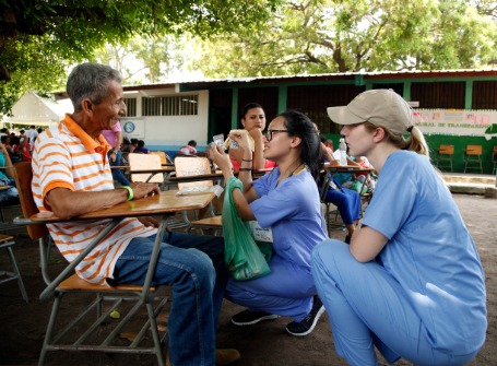 two young women wearing blue scrubs kneel down to talk to a man sitting in a chair