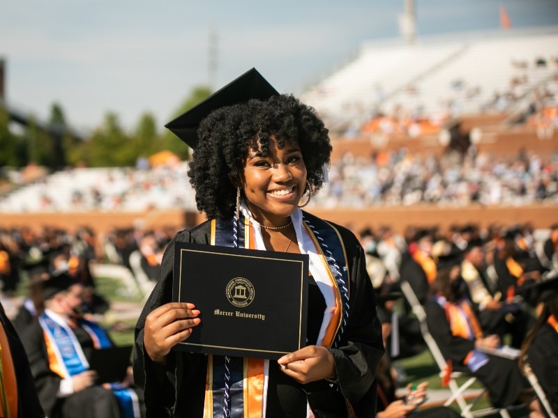 A young woman wearing a cap and gown holds a Mercer degree