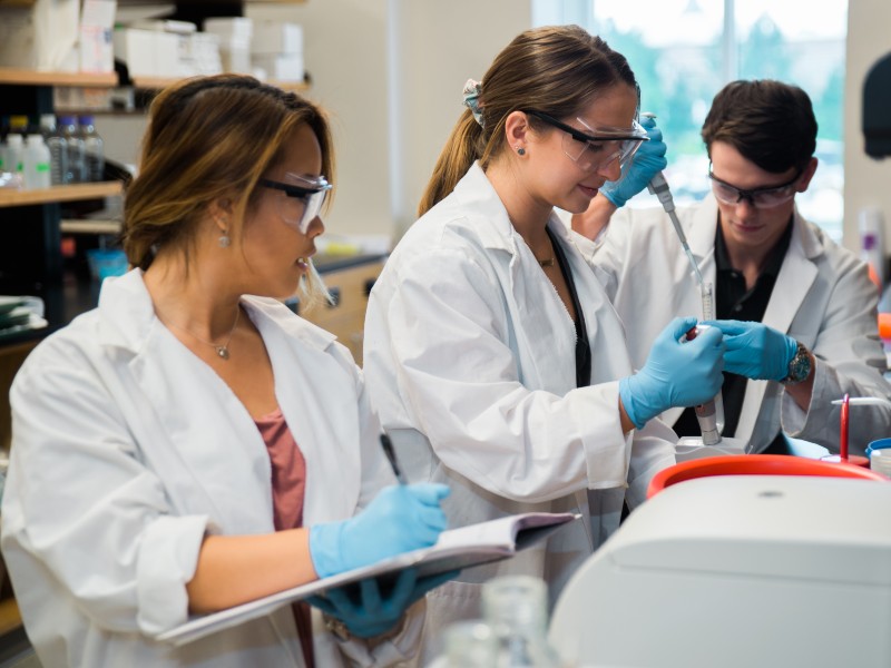Three college students wearing white lab coats work in a lab