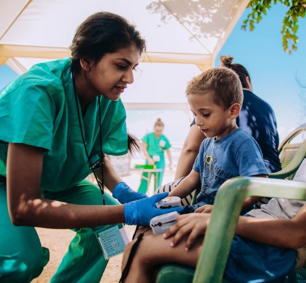 A young woman wearing green scrubs and blue latex gloves checks the pulse ox of a child