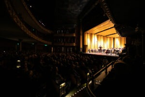 An audience in a darkened concert hall watches a symphony perform on stage
