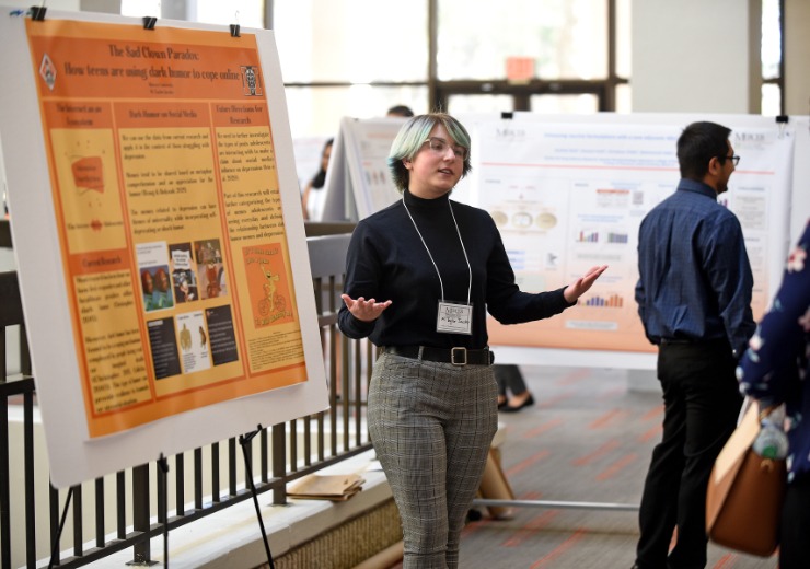 a college student gestures while standing in front of a poster that displays her research