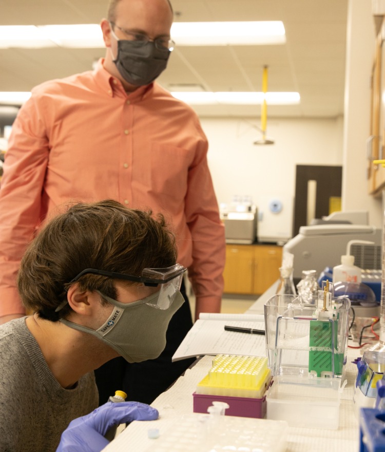 A college student looks closely at chemicals in a lab while a professor watches