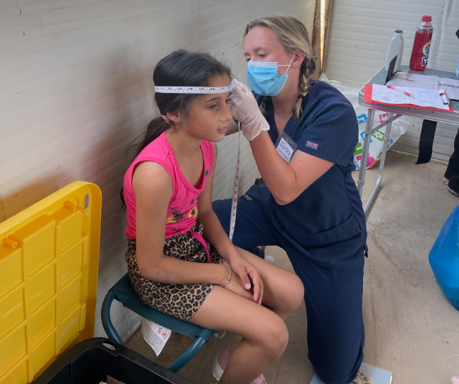 A female student in blue scrubs measures the head circumference of a child