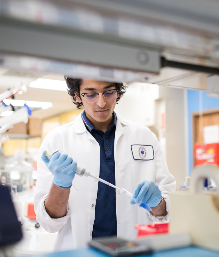 A male college student wearing a white lab coat and blue gloves holds a syringe to the top of a test tube