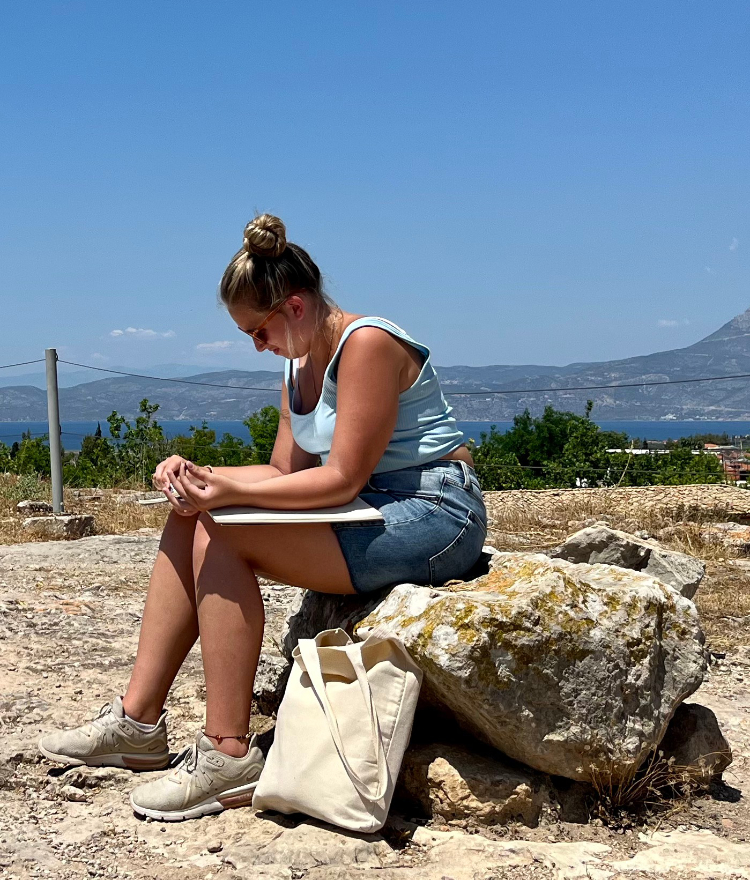 A female college student sits on a rock with a sketchbook open on her lap