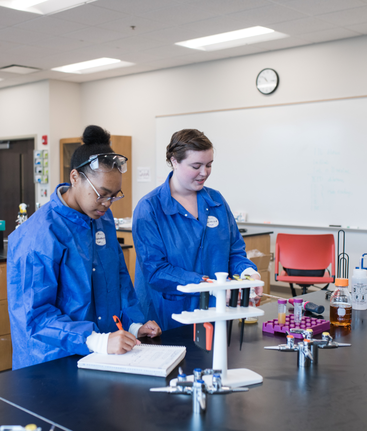 Two female college students wearing blue lab coats work in a laboratory. One is writing in a notebook and the other is touching a beaker.