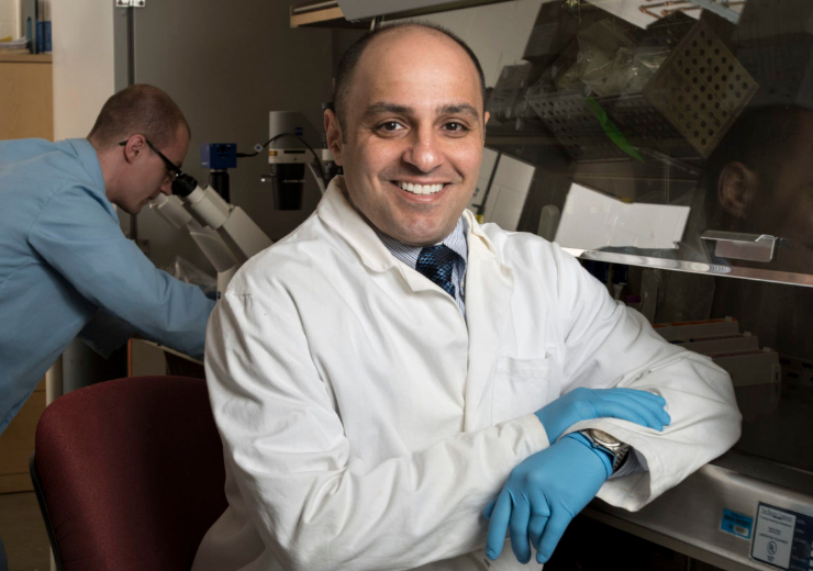 dr. nader moniri sits, posing for a photo wearing a white lab coat and blue latex gloves.