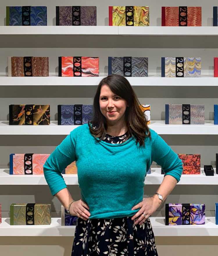 Tennille Shuster stands in front of shelves holding the pandemic journals she handcrafted.