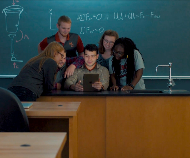 students gather in front of a chalkboard that has equations written on it