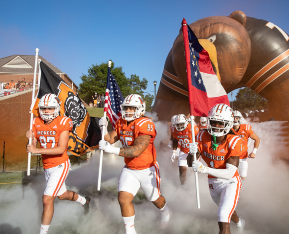 three football players run on the field carrying a mercer flag, american flag and georgia flag