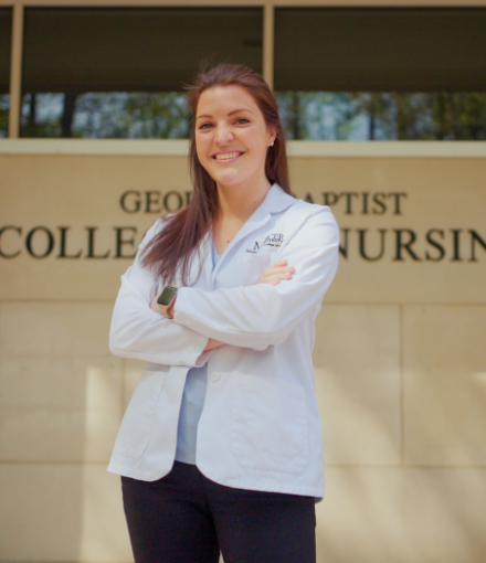 a young woman in a white medical coat stands in front of a sign that says College of Nursing