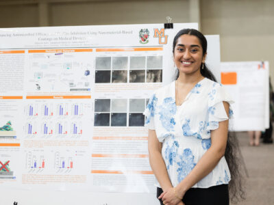 a woman stands in front of a poster titled, "enhancing antimicrobial efficacy and biofilm inhibition using nanomaterial-based coatings on medical devices"