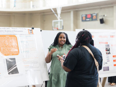 a woman standing in front of a poster talks animatedly to another woman