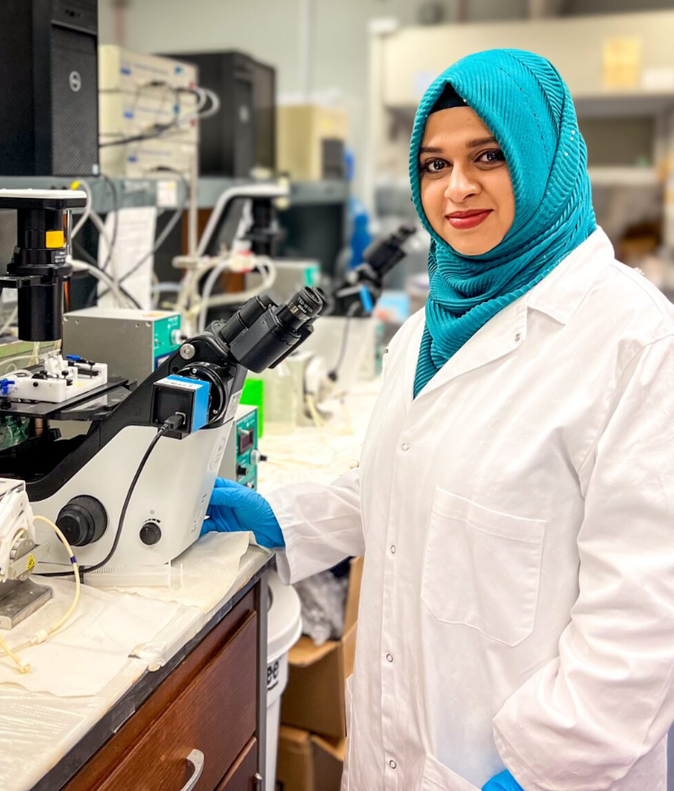 A Mercer postdoctoral fellow stands next to a microscope in a lab.