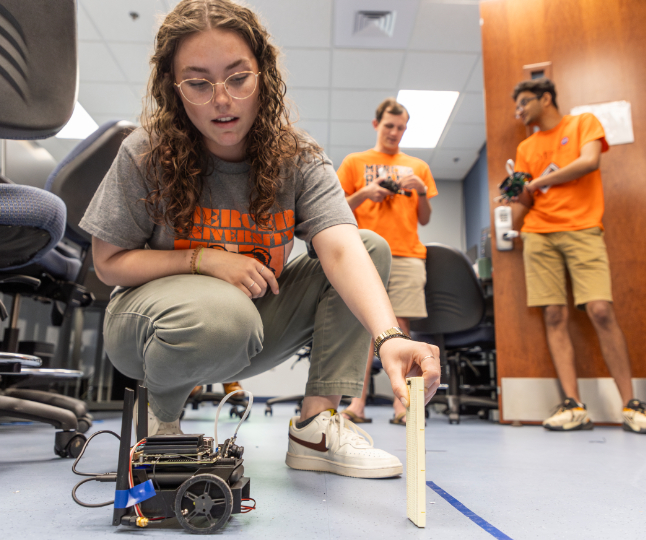 A Mercer University student crouches on the floor of a classroom to work with a small robot.