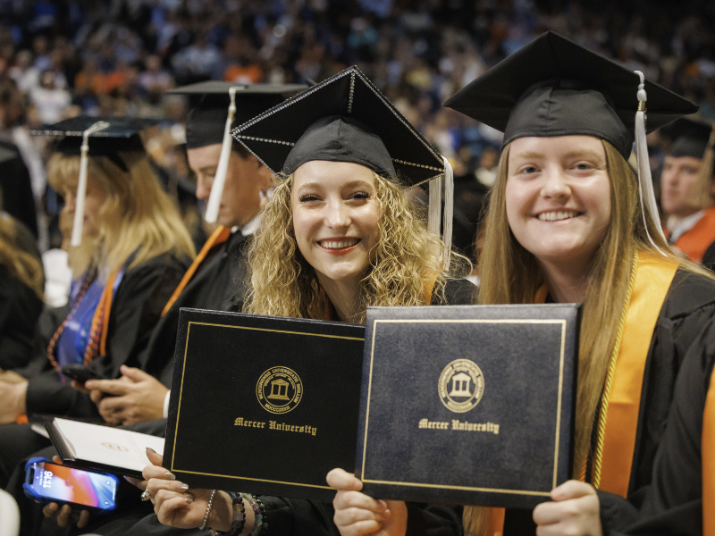 Two graduates in caps and gowns smile and hold diplomas at a Mercer University graduation ceremony.