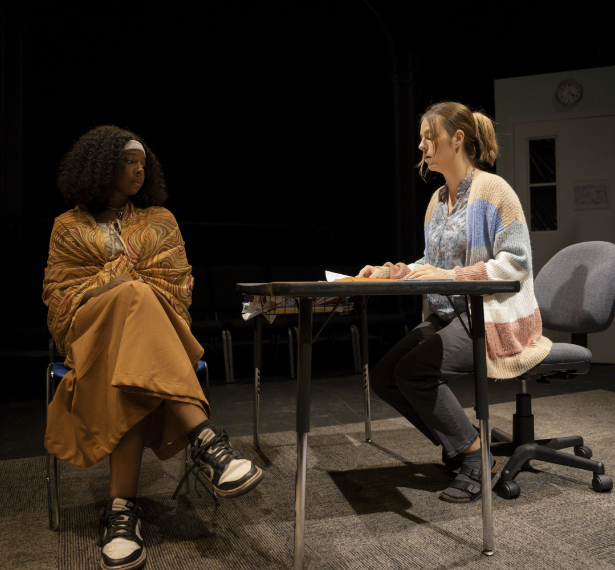 Two women sit across from each other at a table on stage, engaged in conversation under stage lighting.