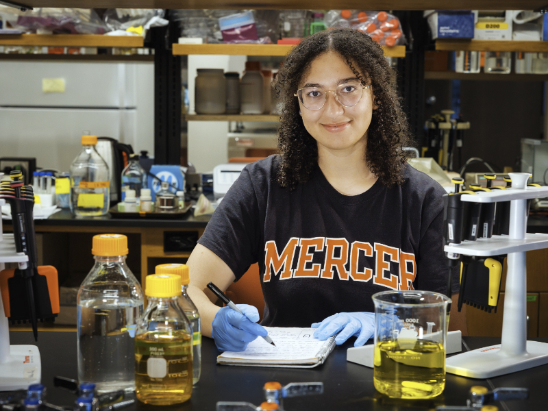 A person wearing gloves and a Mercer shirt writes notes at a lab table with bottles, beakers, and pipettes.
