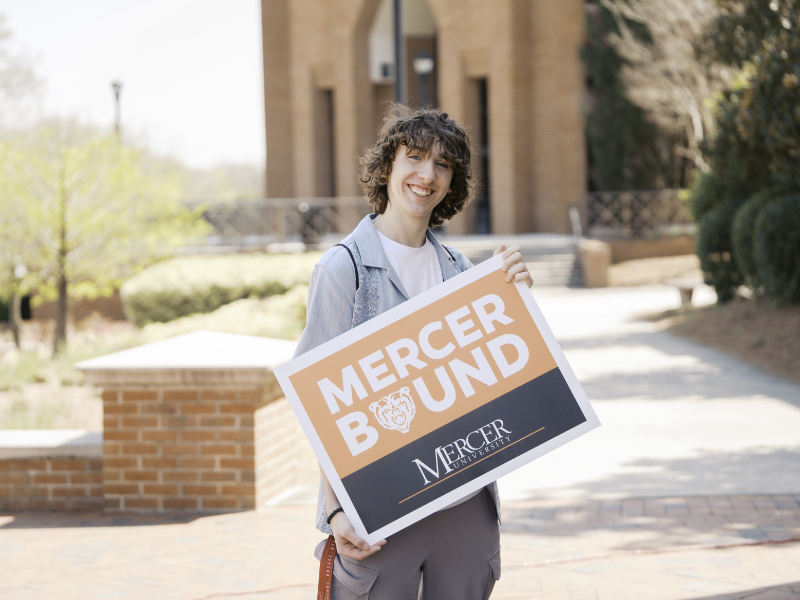 Young man holding a Mercer Bound sign on a brick walkway with a building and trees in the background.