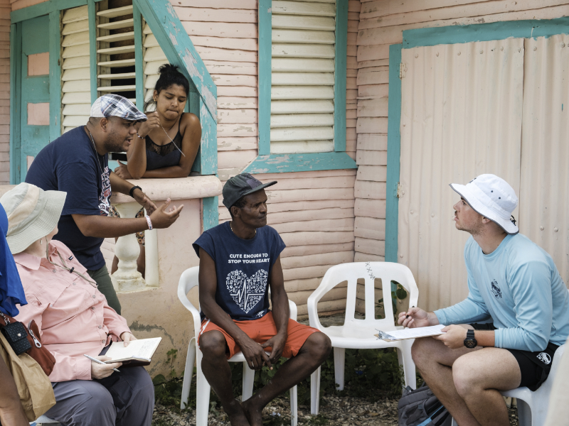 Four people sit and talk outside a pink house while one woman listens from the window; two take notes on clipboards.