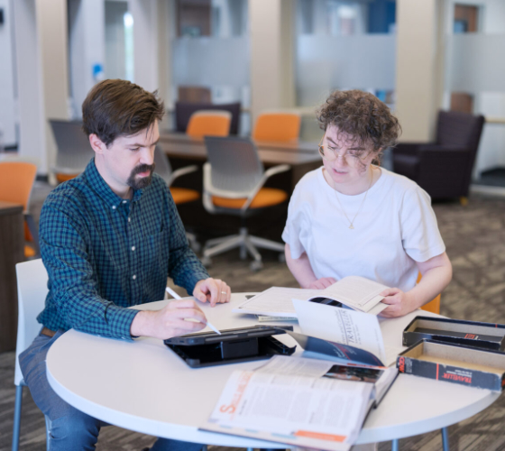 Two people sit at a round table in an office, reading books and taking notes together.