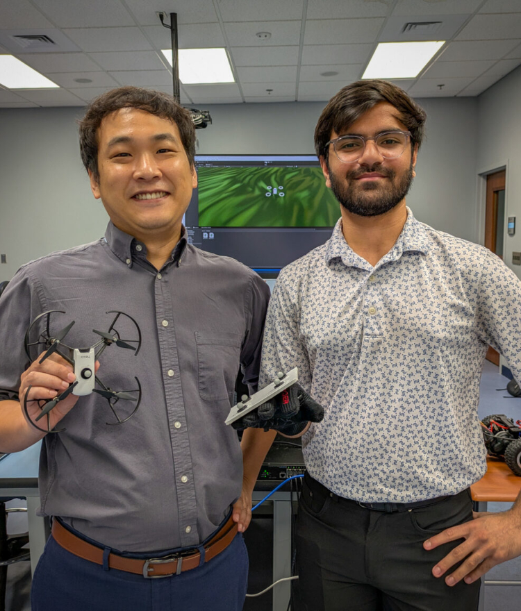 Dr. Hunmin Kim and Shrey Patel in a lab. Dr. Kim holds a drone, and Patel wears a glove with probes affixed to it.