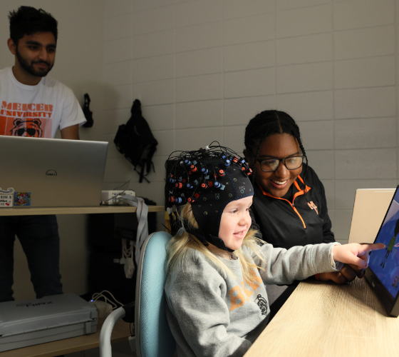 Parth Patel stands at a computer, while Lynzi Holland sits at a table with a child wearing a cap with the fNIRS technology.