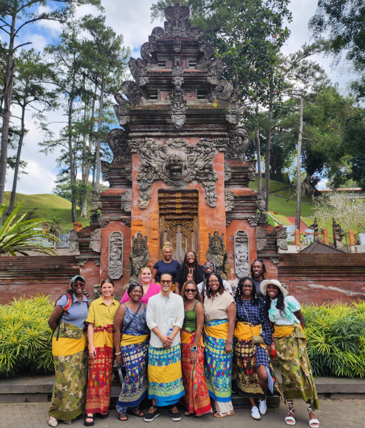 A group of people in colorful sarongs stand in front of an intricately carved stone gate with greenery in the background.