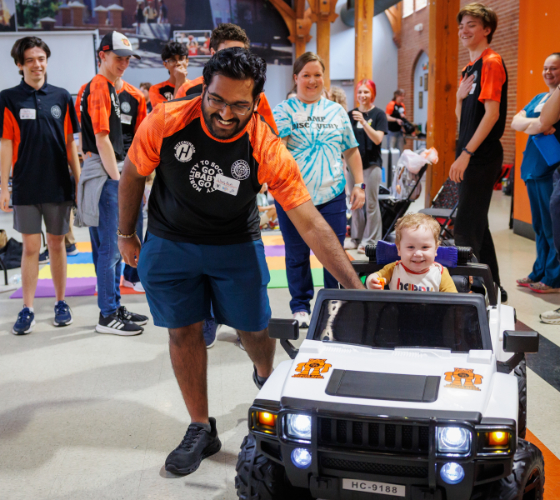 A Mercer student guides a small child riding a toy car, surrounded by smiling people in Penfield Hall.