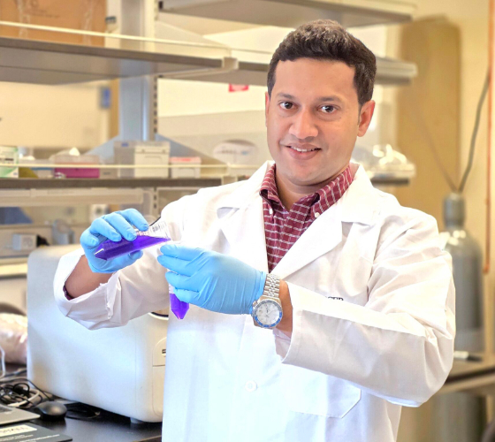 Dr. Raquibul Hasan in a lab coat and gloves handles a test tube with blue liquid in a laboratory setting.