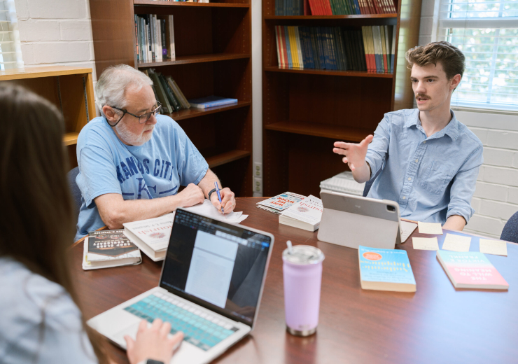 Three people sit at a table with laptops and books, engaged in discussion.