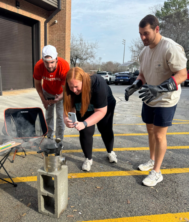 Three people observe and record an experiment with a smoking container on cinder blocks in a parking lot.