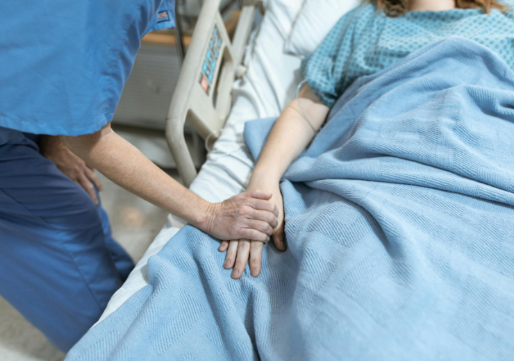 A health care worker holds the hand of a patient lying in a hospital bed covered with a blue blanket.