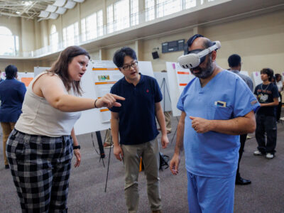 A man wears a virtual reality headset while two students explain something to him.
