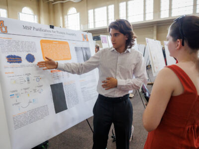 A Mercer student gestures to his poster titled MSP Purification for Protein Assembly while another student looks on.