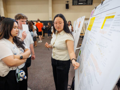 A Mercer student points at her research poster board while two others look on.