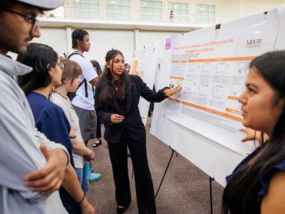 Two Mercer students explain their research poster titled Food Restriction Results in Sex Differences for Palatable Food Seeking to a crowd of people.