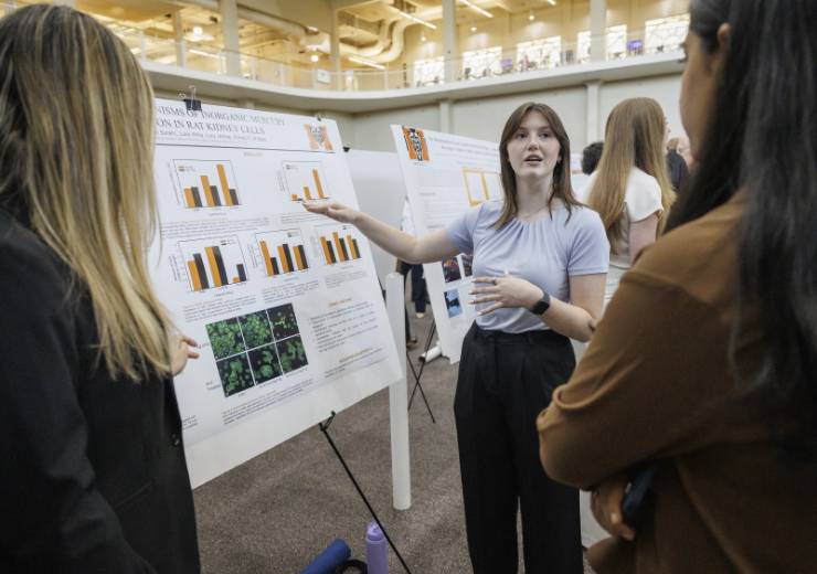 A student presents her research poster on inorganic mercury and rat kidney cells to two attendees at an academic research symposium.