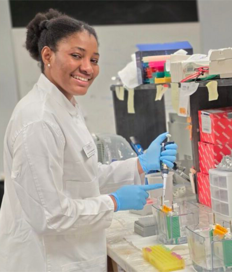 A smiling researcher in a white lab coat and blue gloves uses a pipette at a lab bench stocked with supplies and test tubes.