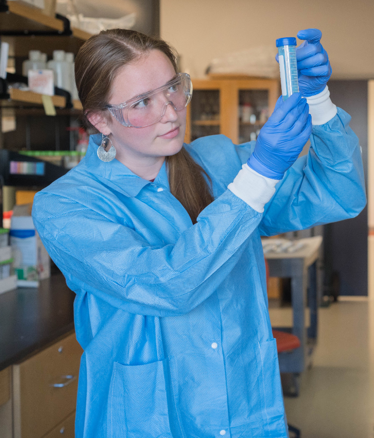 A Mercer student in a blue lab coat, safety goggles, and gloves carefully examines a test tube in a science laboratory.