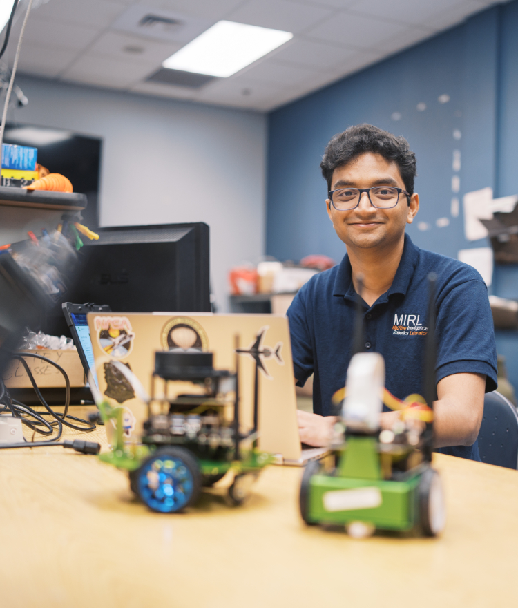 A Mercer student smiles at a desk with small wheeled robots and a laptop in an engineering lab.