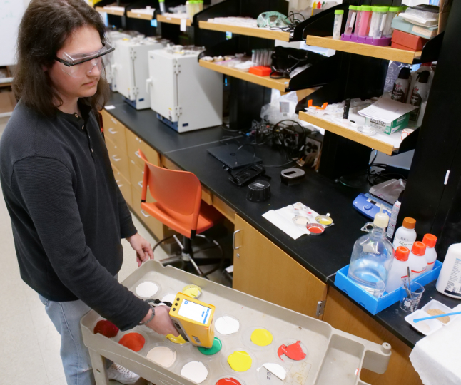 A lab technician wearing safety goggles uses a handheld scanner to test colorful paint samples on a tray in a science laboratory.