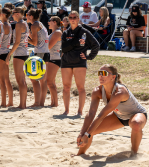 A Mercer beach volleyball player digs for the ball in the sand while teammates and a coach watch from the sideline.