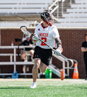 Mercer lacrosse player #2 in a white and black uniform runs with the ball in his stick during a game on the field.