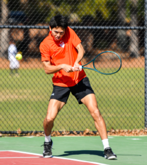 A Mercer University men's tennis player in an orange shirt hits a backhand shot during an outdoor match.
