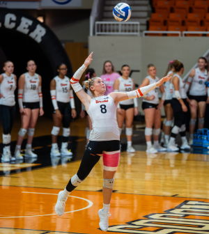 Mercer volleyball player #8 serves the ball during a home match with teammates lined up in the background.