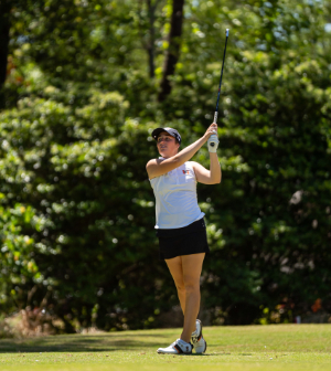 A Mercer University women's golfer follows through on a swing on a tree-lined course during a sunny tournament round.